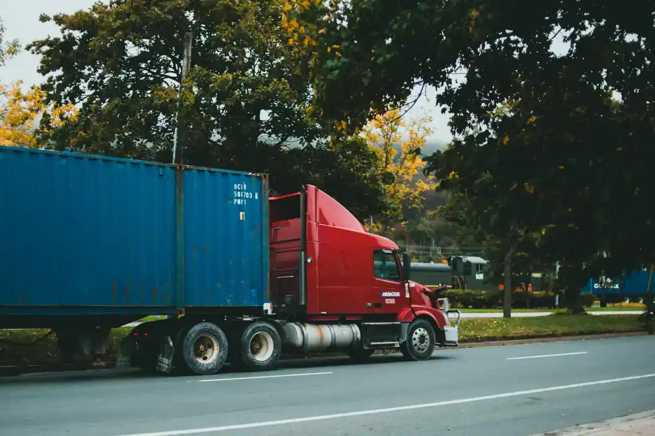 Red semi truck hauling a blue shipping container down a tree-lined road